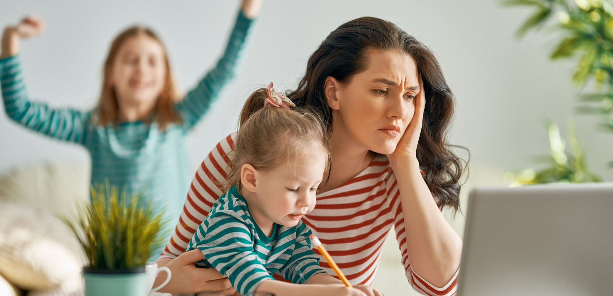 Frau mit zwei kleinen Kindern vor dem Laptop mit gestresstem Blick.